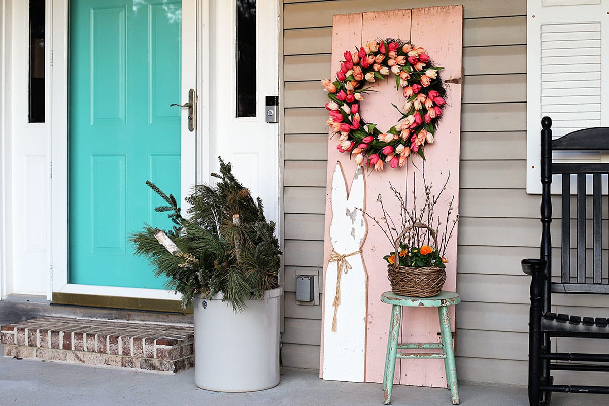 Spring front porch with a pink wooden door, colorful tulip wreath, wooden Easter bunny and a basket of yellow pansies.