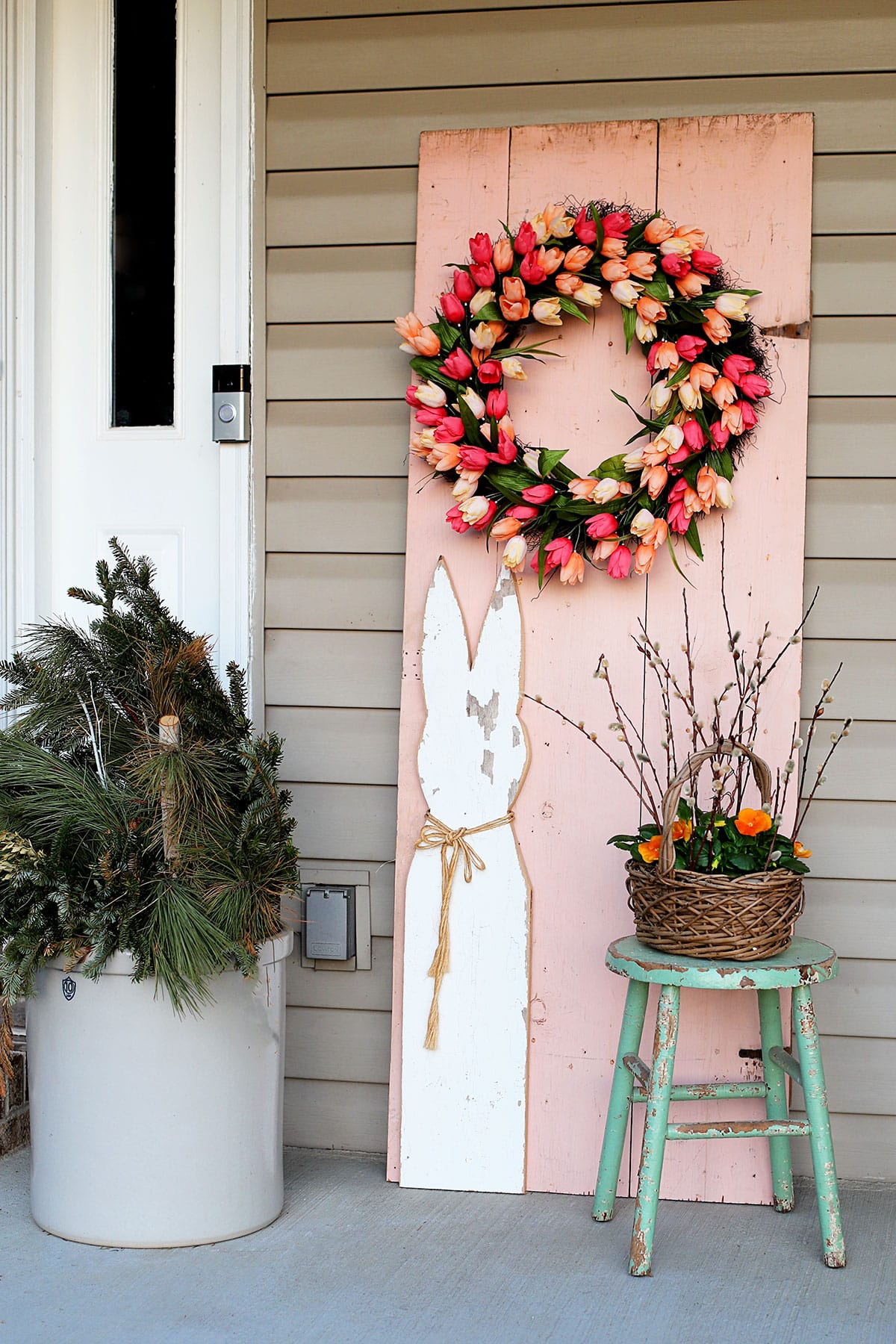 Spring front porch with a pink wooden door, colorful tulip wreath, wooden Easter bunny and a basket of yellow pansies.